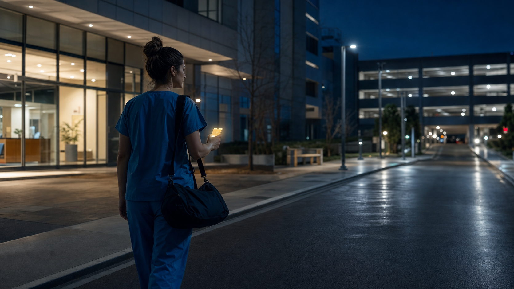 A nurse leaving a hospital after a night shift, walking toward a distant parking structure with a subtle phone safety glow